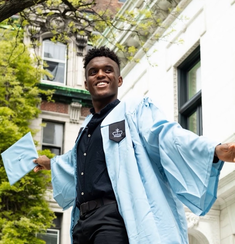 Omar at graduation in a light-blue Columbia gown, arms out, grinning, cap in one hand.